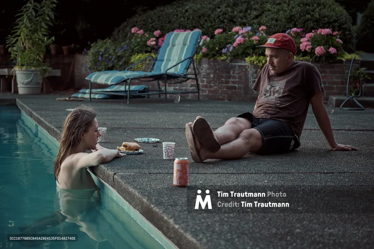 At dusk, a woman with wet hair rests her arms on the edge of a backyard swimming pool, chatting with a man who sits on the pool deck beside her. Between them sit plates of food, patterned paper cups, and a canned drink. The man wears a red baseball cap, a brown t-shirt, and dark shorts. A turquoise lounge chair, brick retaining wall, and lush pink flowering shrubs are visible in the softly lit background.