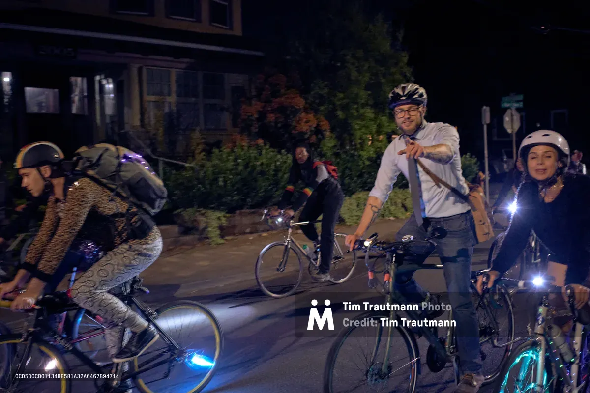 A spirited group of cyclists assembles on Southeast César E. Chávez Boulevard during Portland's Drop Out Prom Ride, part of the city's celebrated Pedalpalooza festival in June 2019. LED lights pierce the darkness as helmeted riders of various ages pause on the residential street, their bikes illuminated by colorful safety lights. The nocturnal scene captures the grassroots energy of Portland's cycling community, with mature trees and craftsman-style homes creating an intimate neighborhood backdrop for this unconventional prom celebration.