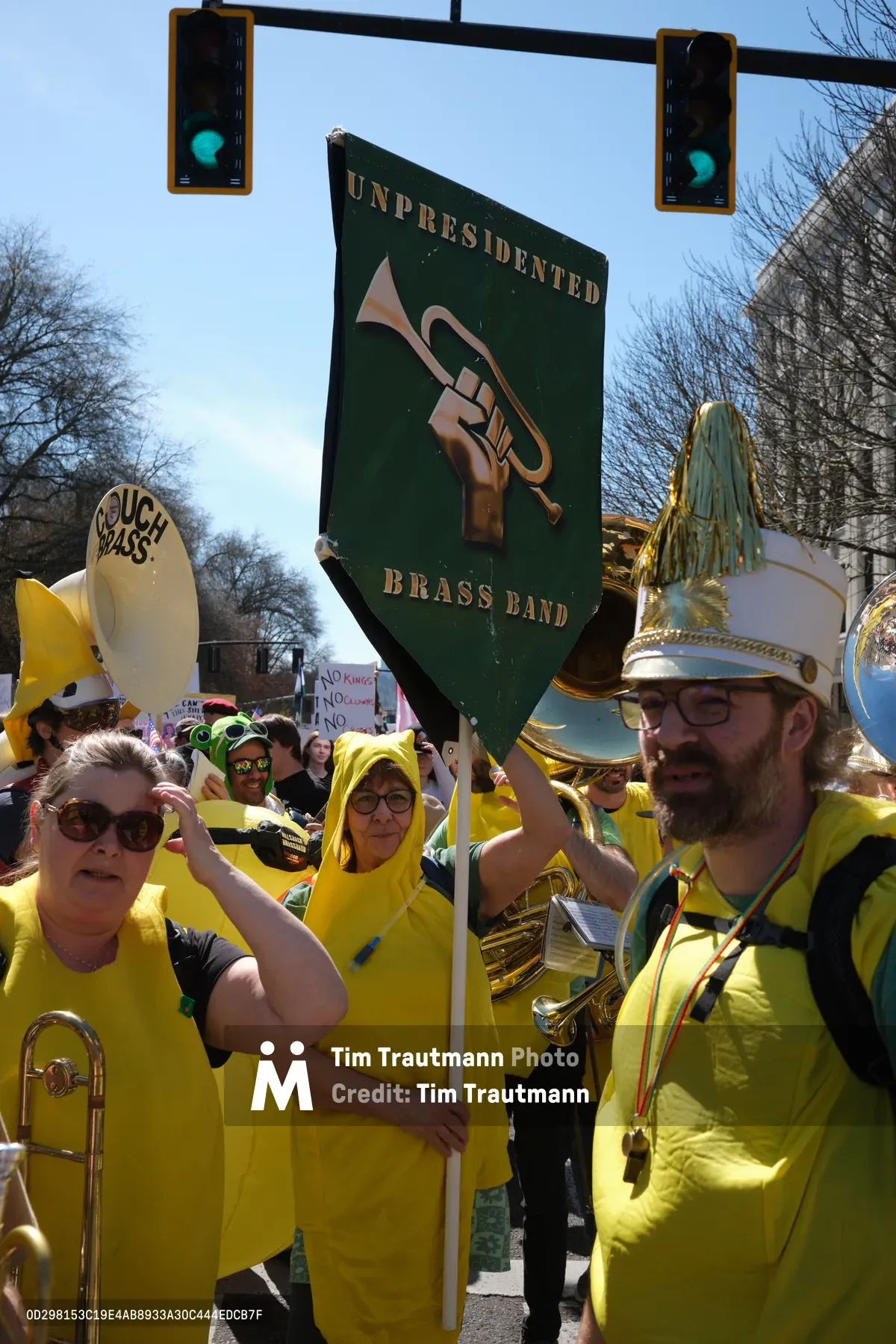 Members of the Unpresidented Brass Band march through Portland's Skidmore/Old Town Historic District during a 2026 'No Kings' demonstration, their vibrant yellow uniforms creating a striking sea of color against the pale spring sky. A bearded musician in a plumed hat raises the band's green banner bearing their emblematic hand-and-trumpet logo, while fellow protesters clutch brass instruments and signs beneath dual green traffic lights. The scene captures the spirited intersection of musical activism and political resistance on Southwest Ankeny Street.