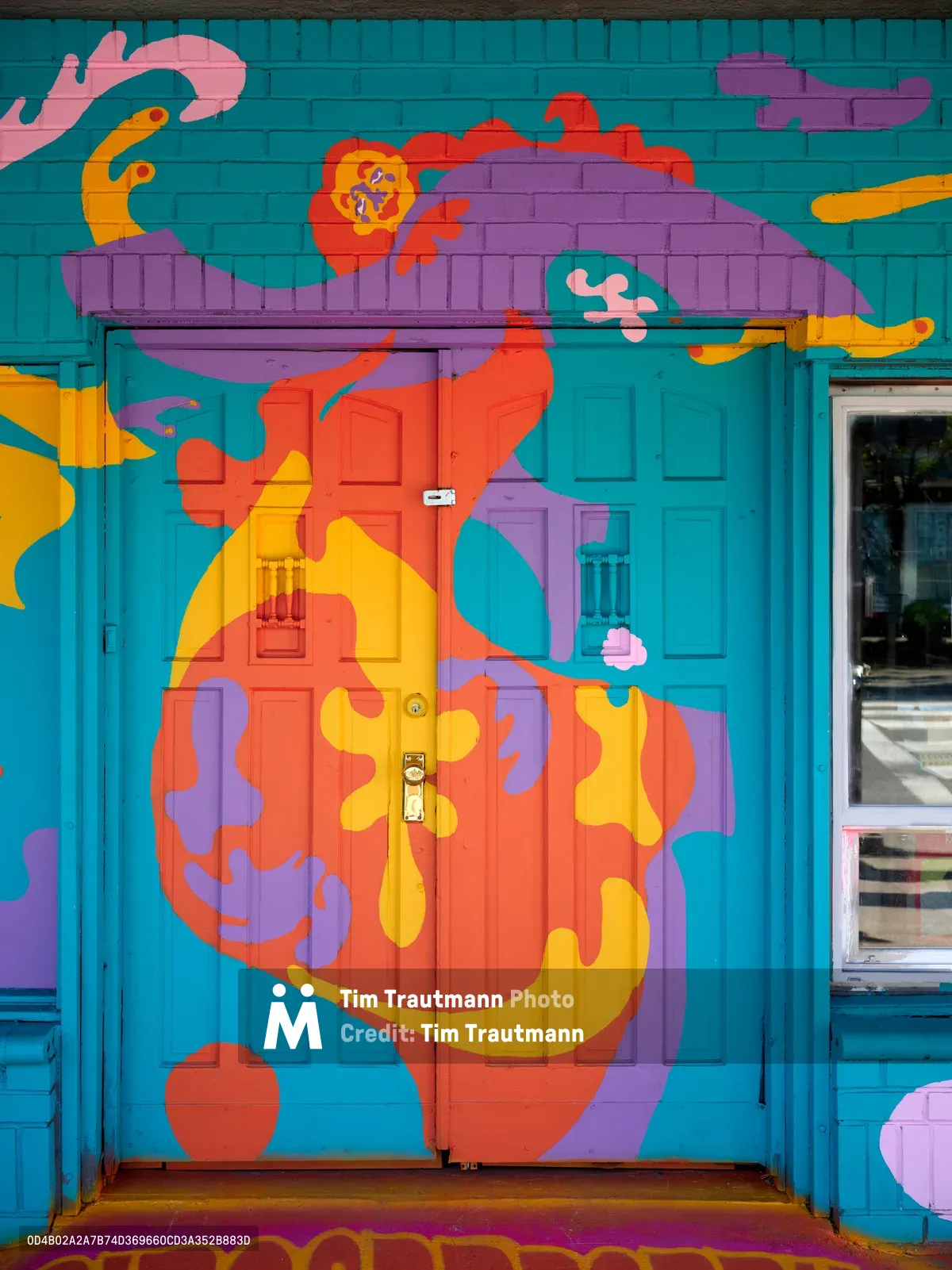 An abstract human figure dominates this explosive street mural by Naomi Likayi, painted across turquoise wooden doors and brick walls in Portland's Alberta Arts District. The character is adorned in flowing orange and purple garments, with wild hair rendered in kaleidoscopic hues against the cerulean backdrop. Bold geometric shapes and organic forms dance around the central figure, transforming an ordinary storefront entrance into a portal of artistic fantasy and neighborhood creative expression.