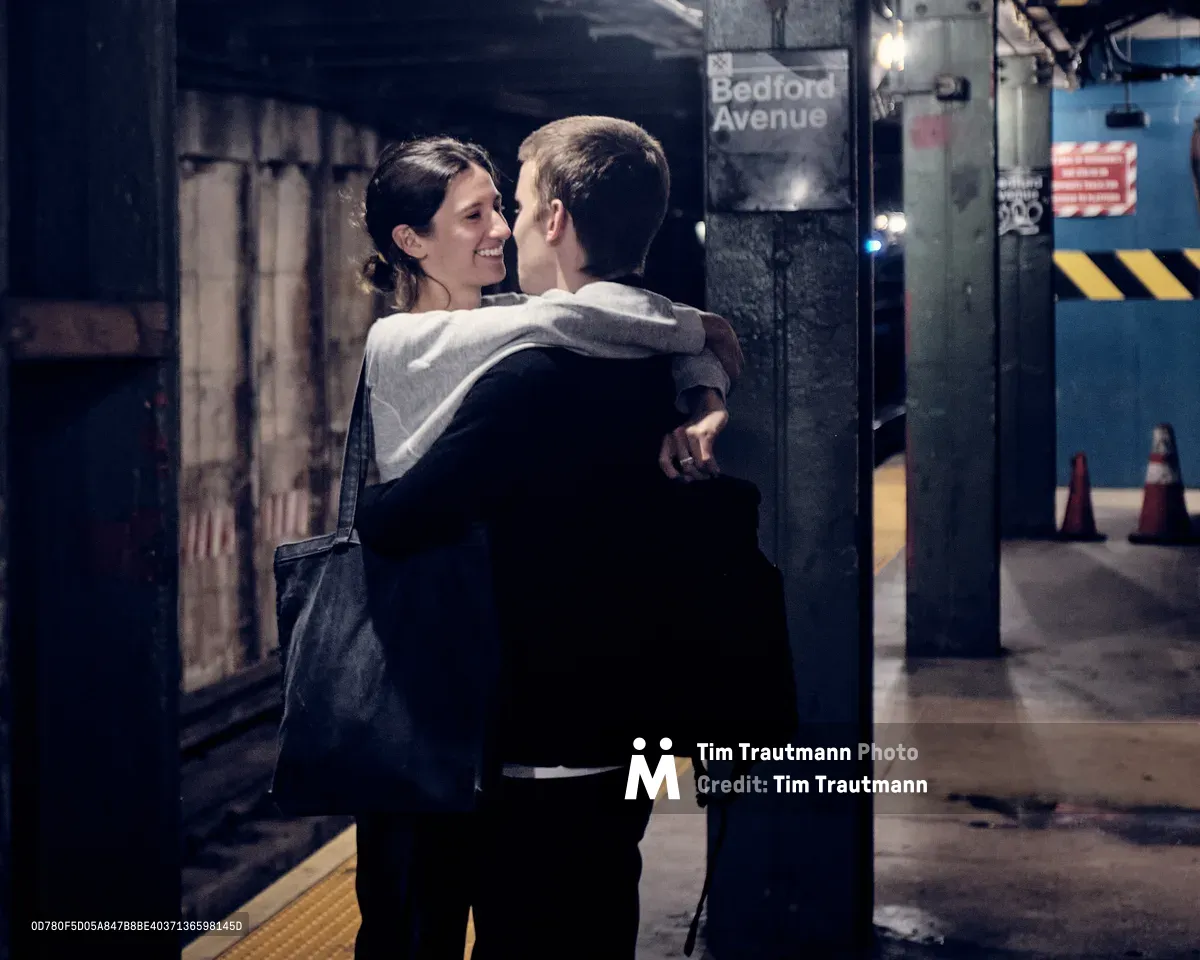A tender moment unfolds on the platform of Bedford Avenue subway station in Williamsburg, Brooklyn, as a couple shares an intimate embrace beneath the station's iconic signage. The woman, wearing a light hoodie and carrying a dark bag, smiles warmly as she holds her partner dressed in dark clothing. The scene captures the raw authenticity of New York subway romance, with the station's industrial elements—weathered tiles, warning signs, and platform infrastructure—creating a gritty urban backdrop bathed in the station's harsh fluorescent lighting.