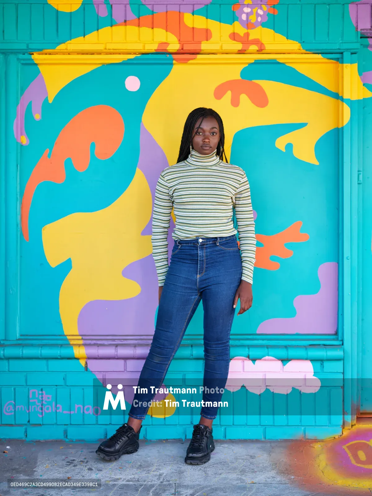 Naomi Likayi stands confidently before her completed mural on Northeast 15th Avenue in Portland's Alberta district. The young Black artist wears a striped turtleneck and high-waisted jeans against the explosive backdrop of turquoise, yellow, orange, and purple abstract shapes that dance across the storefront's surface. Natural daylight illuminates both subject and artwork, creating a harmonious composition that celebrates urban art and creative expression in this gentrifying Portland neighborhood.