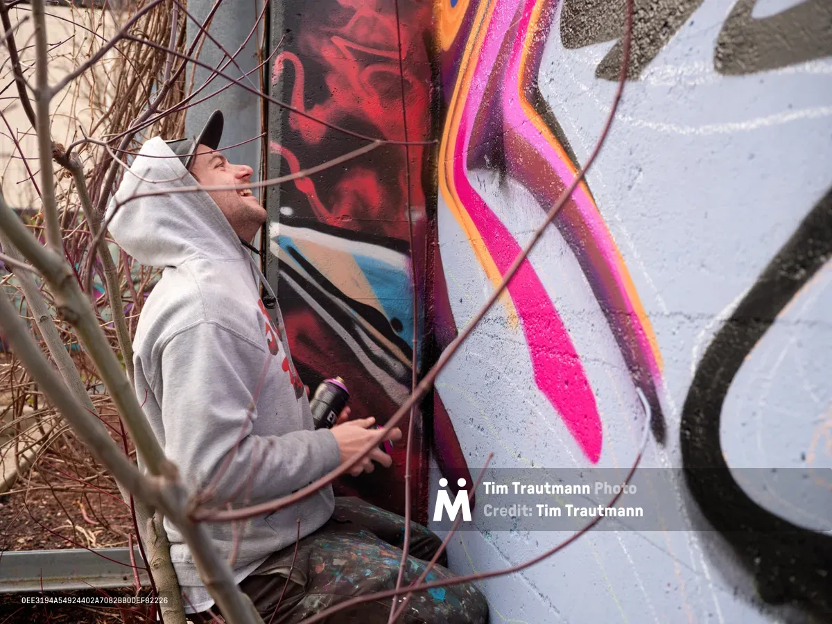Travis Fields, known as Campographic, crouches beside a concrete wall at Clay Creative Building in southeast Portland's Hosford-Abernethy neighborhood, spray can in hand as he works on a bold mural. The artist, wearing a light gray hoodie, gazes upward at his evolving creation of flowing magenta, orange, and crimson forms against weathered concrete. Dormant winter grasses frame the intimate creative moment, capturing the meditative focus of urban artistry in progress.