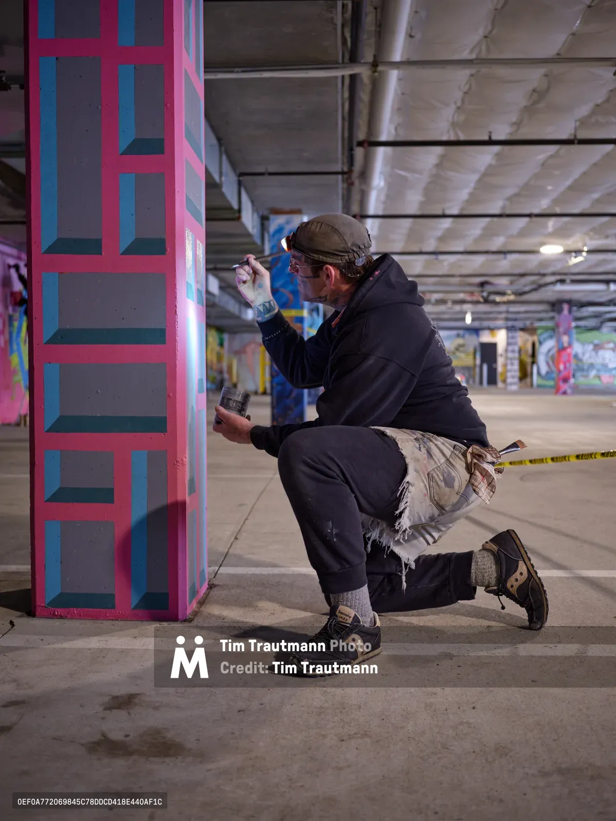 Bike activist and artist Rabbitt kneels in focused concentration as he applies precise brushstrokes to a vibrant geometric mural on a concrete column in the Clay Creative building's parking garage. The underground space, illuminated by harsh fluorescent lighting, transforms into an impromptu studio where pink and blue angular patterns emerge against the industrial backdrop. His paint-stained work clothes and methodical approach capture the dedication required to bring color to Portland's urban spaces.