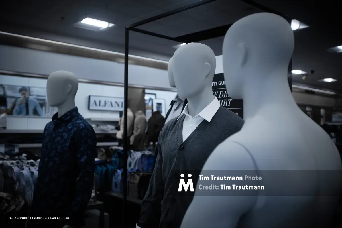 Three mannequins stand in stark contrast within Nordstrom's Clackamas location, their featureless forms creating an unsettling tableau under harsh fluorescent lighting. Two figures are impeccably dressed in professional attire—one in a navy patterned shirt, another in a charcoal vest with crisp white collar—while a third stands naked, its pale form catching the light like an uninvited guest at a formal gathering. The scene captures the surreal intersection of commerce and humanity, where plastic bodies serve as silent ambassadors for fashion consumption.