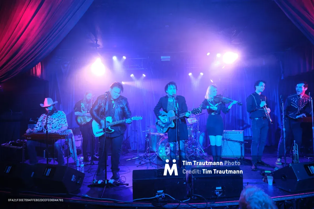 Six-piece band Roselit Bone performs under dramatic blue and purple stage lighting at the intimate Dante's venue in Portland, Oregon during RX Fest 2019. The atmospheric concert photography captures the musicians mid-performance, with the lead vocalist at center microphone flanked by guitarists, a violinist, and a keyboardist in cowboy hat. Theatrical lighting cuts through hazy air, creating a moody underground club aesthetic framed by burgundy curtains.