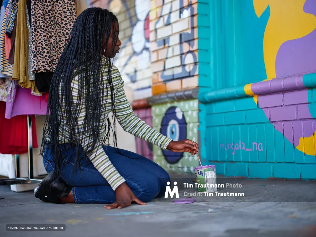 Naomi Likayi kneels on concrete pavement as she carefully signs her vibrant mural on a turquoise and purple brick wall in Portland's Alberta neighborhood. Her long braided hair falls forward as she concentrates on her brushwork, wearing a striped long-sleeve shirt and blue jeans. The colorful street art backdrop includes clothing displays and urban elements that speak to the area's creative energy. Warm natural light illuminates this moment of artistic completion, capturing the intimate act of an artist claiming her work.