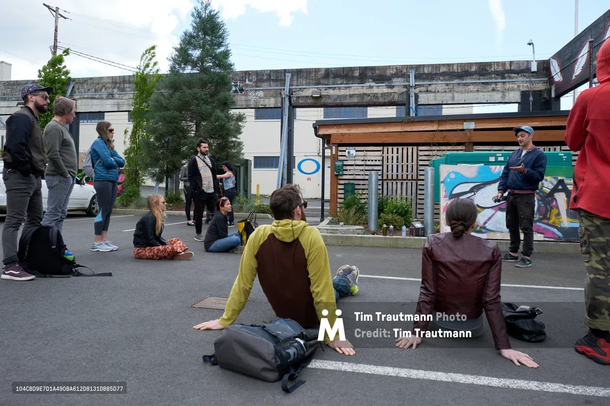 Galen Malcolm of the Portland Street Art Alliance captivates a diverse group of tour participants with a live spray painting demonstration outside Clay Creative in Portland's Hosford-Abernethy neighborhood. The intimate gathering sits and stands on the asphalt street, their attention fixed on the artist working at a colorful graffiti wall beneath overcast Pacific Northwest skies. The industrial backdrop of concrete buildings and power lines frames this grassroots cultural exchange, where street art education meets community engagement in Southeast Portland's creative corridor.