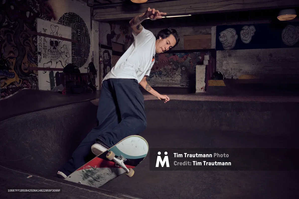 A tattooed skateboarder in white shirt and dark pants carves through a concrete bowl at Commonwealth Skateboarding in Portland's Buckman neighborhood. The underground venue's raw industrial atmosphere is punctuated by graffiti-covered walls, exposed beams, and skull artwork, creating a gritty backdrop for this editorial skateboarding session. Warm lighting illuminates the rider's focused expression as he navigates the curved concrete surface.