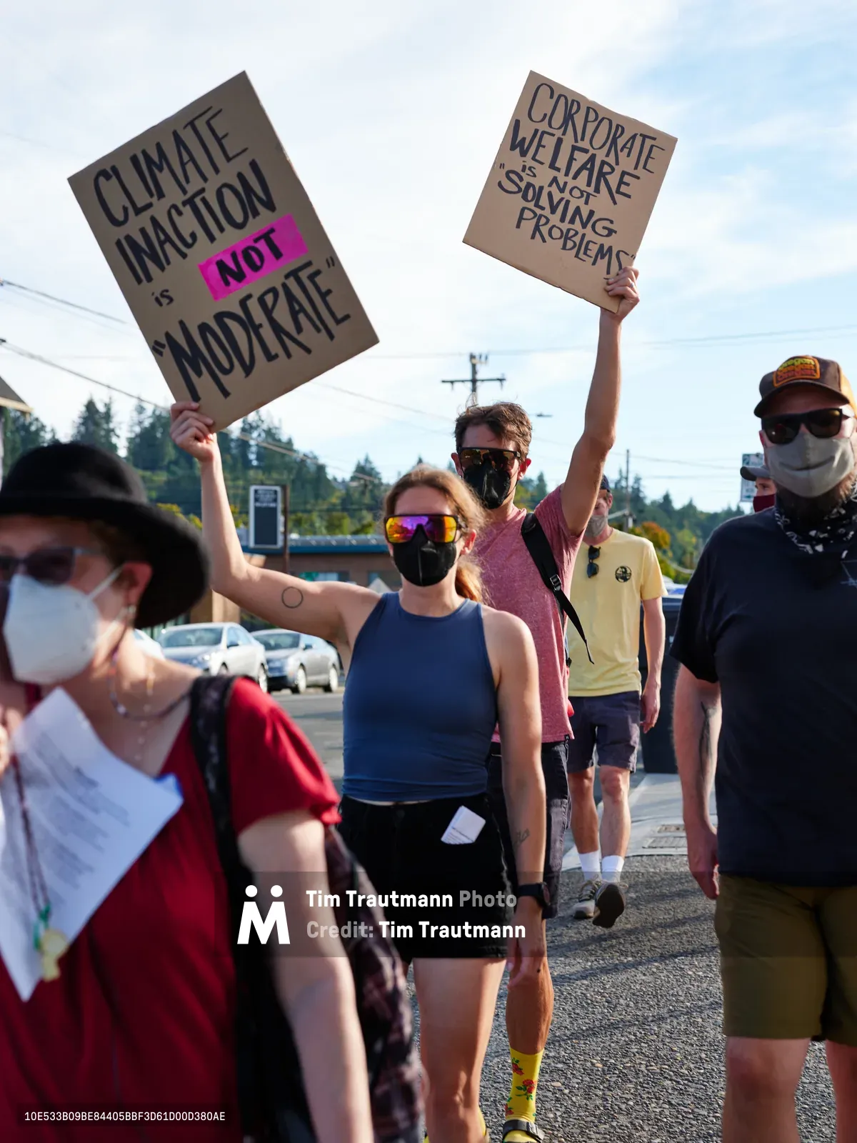 Masked protesters stride purposefully down McLoughlin Promenade in Oregon City, their handmade cardboard signs declaring "Climate Inaction is NOT Moderate" and "Corporate Welfare Not Solving Problems" against a backdrop of power lines and suburban storefronts. The afternoon sun casts sharp shadows across the asphalt as the determined group advances toward Representative Kurt Schrader's office. Their reflective sunglasses and face coverings create an anonymous solidarity while colorful athletic wear adds vibrancy to this grassroots demonstration for environmental action.
