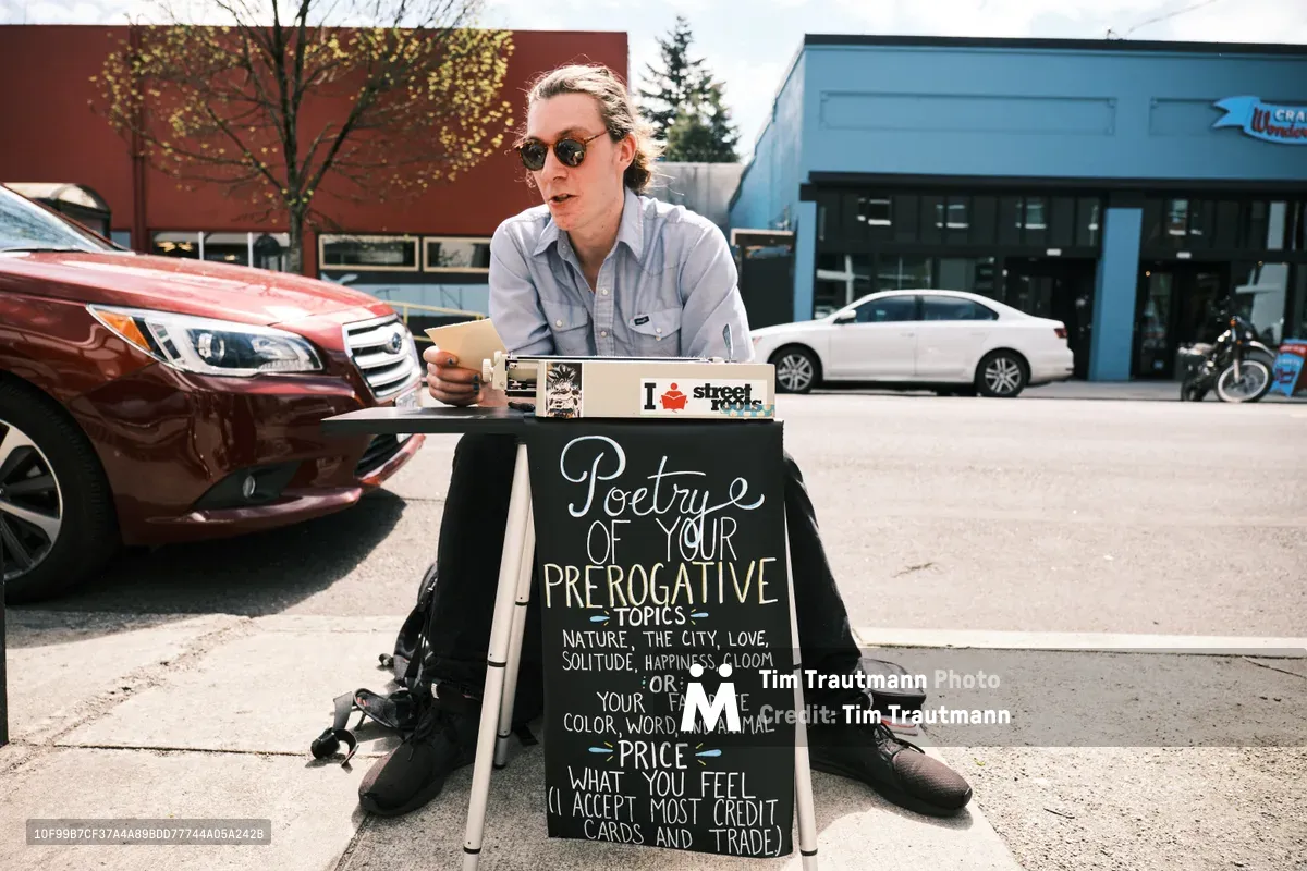 Till Gwinn, a local street poet, sits with legs spread apart behind his vintage typewriter on Portland's Alberta Street, putting finishing touches on a custom poem for a customer. His chalkboard sign advertises 'Poetry of Your Prerogative Topics' with handwritten details about pricing and payment options. The afternoon light bathes the scene as cars pass by on this bustling Northeast Portland thoroughfare, capturing the intersection of urban life and creative expression in one of the city's most vibrant neighborhoods.