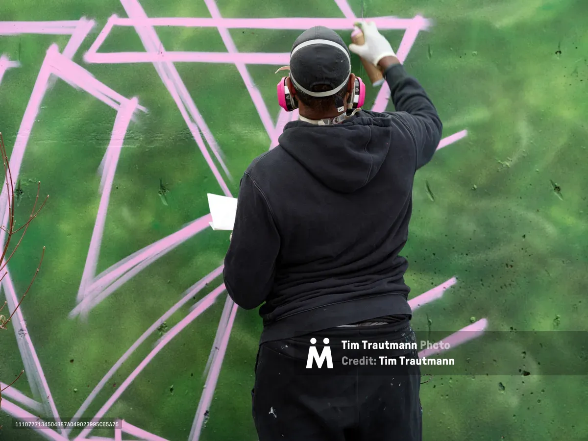 A muralist in black hoodie and baseball cap works intently on a dynamic pink and green graffiti piece at Clay Creative in Southeast Portland's Hosford-Abernethy neighborhood. The artist reaches upward with spray can in hand, adding flowing pink outlines to an emerald green background that dominates the wall surface. Red headphones frame their focused concentration as they craft bold geometric patterns and sweeping letterforms across the urban canvas.