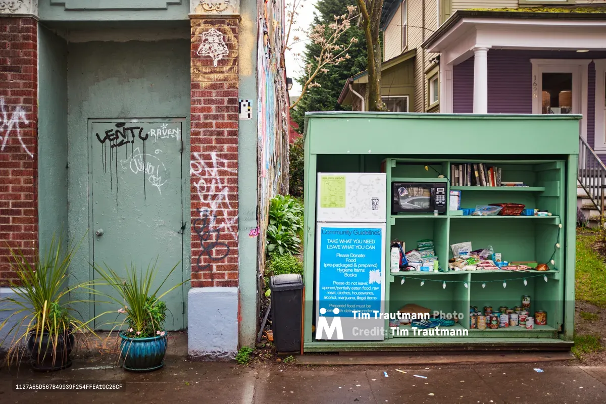 A sage green community fridge stands on Northeast Alberta Street in Portland's Vernon neighborhood, its shelves stocked with canned goods, books, and household items available to anyone in need. The weathered urban setting features graffitied doors, eclectic residential architecture, and potted plants creating an authentic street scene. Blue informational signage explains the mutual aid guidelines while string lights add warmth to this grassroots food security initiative.