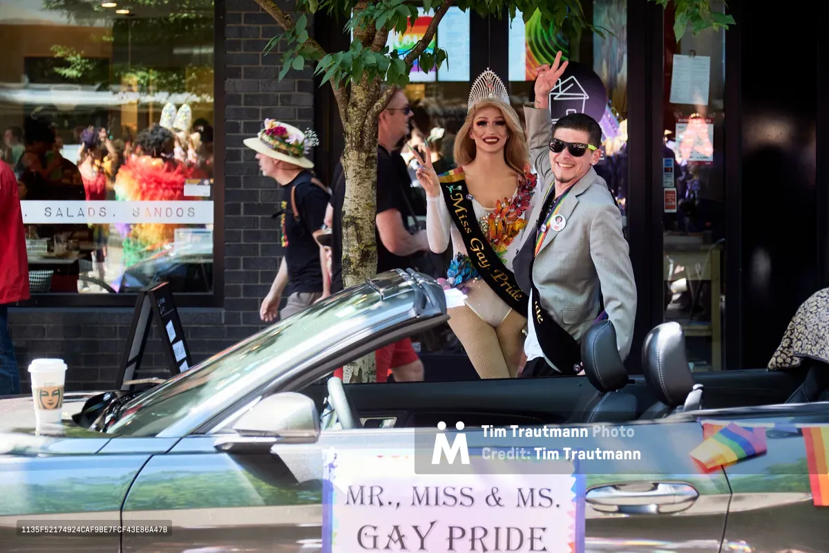 In Portland's Pearl District, a crowned Miss Gay Pride and suited Mister Gay Pride stand beside their silver convertible, preparing for the 2019 Pride Parade. The tiara-wearing queen in her ornate sash and her dapper male counterpart pose with radiant joy before the procession begins, both embodying the celebratory spirit of LGBTQ+ visibility. Dappled sunlight filters through leafy trees onto the busy sidewalk scene, where onlookers gather outside brick storefronts in anticipation of this moment of community pride and representation.