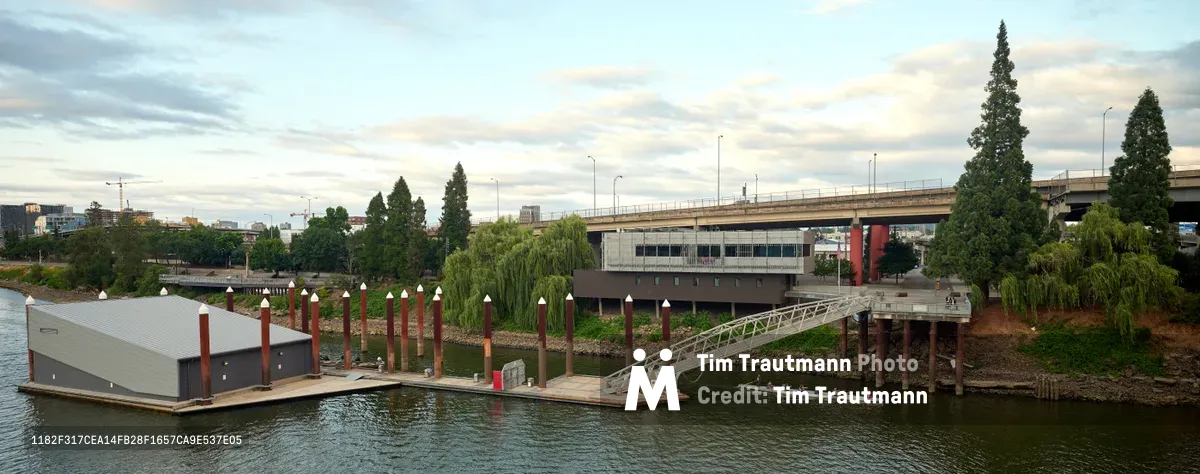 Portland Fire & Rescue Station 21 sits serenely on the Willamette River at the foot of Southeast Madison Street, its modern floating structure anchored by distinctive red pilings that march in formation toward the shore. The corrugated metal boathouse reflects the overcast Pacific Northwest sky while a white pedestrian bridge connects the water-based facility to the Central Eastside embankment. Towering evergreens and urban infrastructure frame this unique emergency response outpost, where tradition meets innovation in river rescue operations.