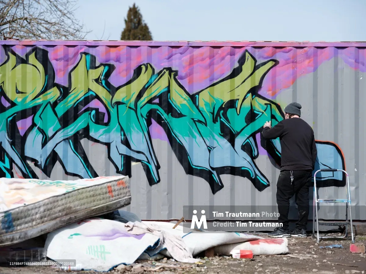 A graffiti artist in black clothing applies finishing touches to a dynamic wildstyle piece featuring flowing turquoise and lime green letters against a purple-pink background. The scene unfolds on a corrugated metal fence in Northeast Portland, Oregon, with scattered paint-stained tarps and a step ladder indicating an active work session. Bare winter trees and overcast skies frame this urban art creation, capturing the raw energy of street culture and creative expression.