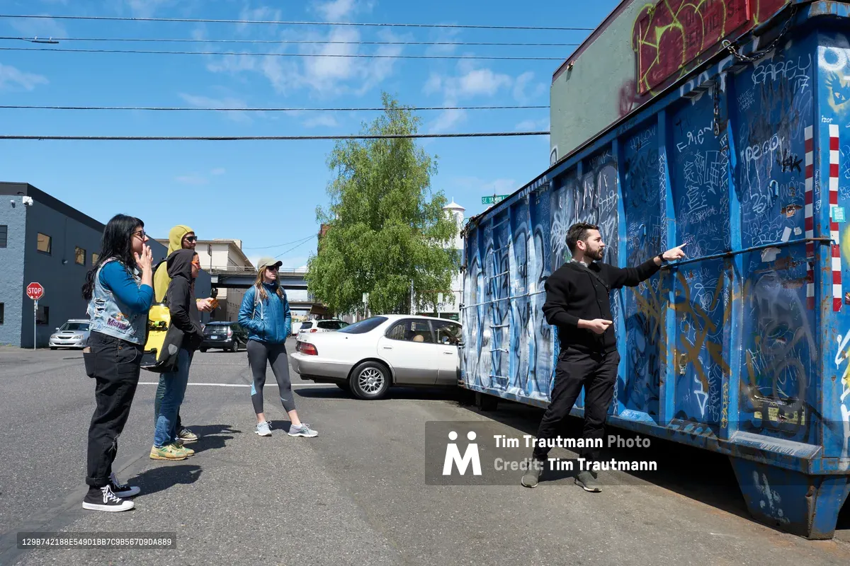 Tomás Valladares of the Portland Street Art Alliance gestures toward vibrant blue graffiti adorning a dumpster while leading an engaged group of young adults through Portland's Central Eastside. The afternoon scene captures the intersection of grassroots cultural education and urban expression, as participants listen attentively against a backdrop of industrial buildings and power lines. Sunlight illuminates the weathered asphalt and eclectic mix of street art that transforms utilitarian infrastructure into canvases of creative rebellion.