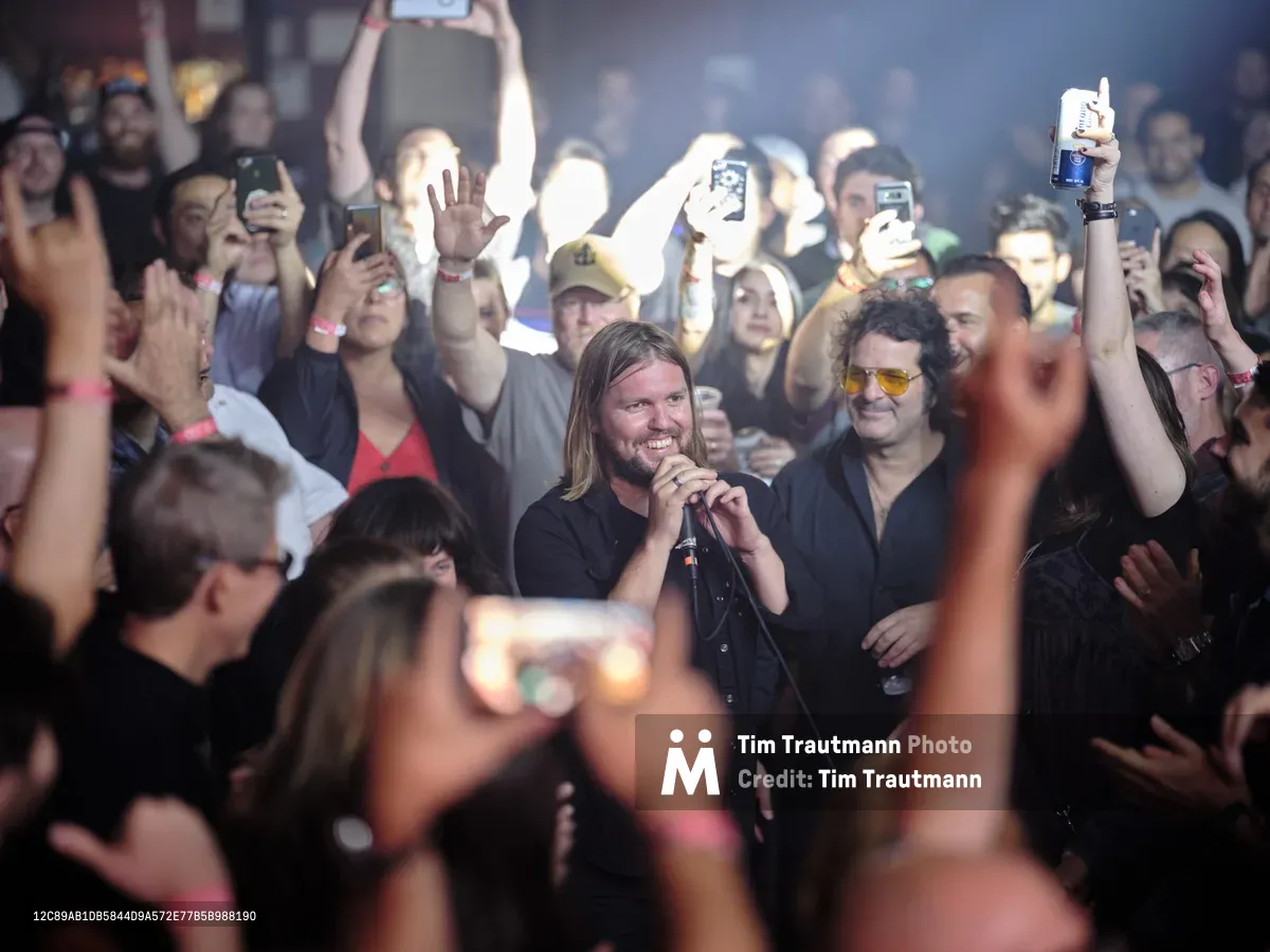 Band of Skulls frontman Russell Marsden grips the microphone with infectious energy as he performs at Webster Hall's intimate stage, surrounded by a sea of glowing smartphone screens held aloft by devoted fans. The atmospheric lighting cuts through the haze, illuminating his long blonde hair and genuine smile while capturing the electric connection between performer and audience. Behind him, bandmate Emma Richardson shares in the moment, while the packed crowd creates a constellation of digital documentation in the historic Manhattan venue.
