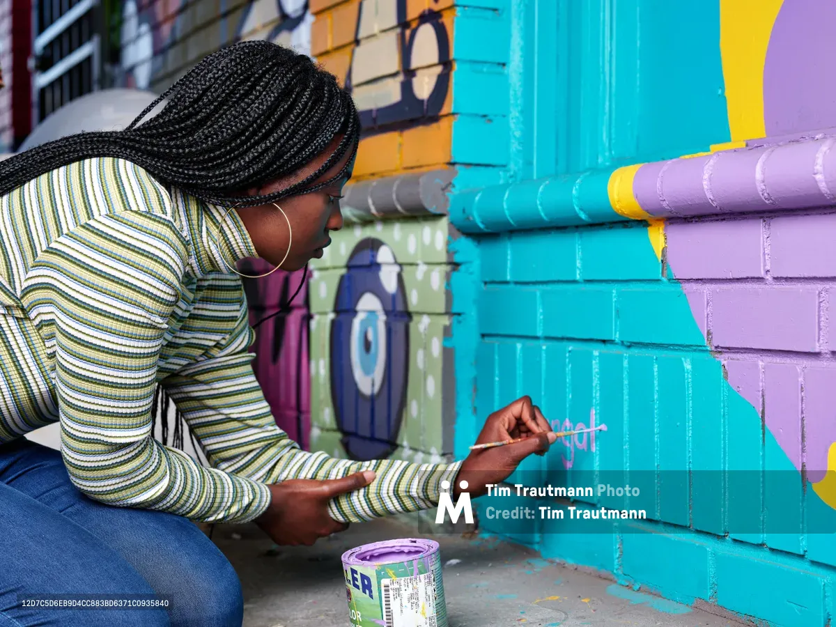 Naomi Likayi kneels beside her vibrant mural on Alberta Street, carefully applying her signature with a fine brush to the electric blue section of the painted brick wall. Her intricately braided hair cascades as she leans into her work, wearing a striped long-sleeve top that echoes the rhythmic patterns of her street art. Behind her, the colorful geometric mural stretches across the wall in bold turquoise, purple, and yellow blocks, transforming the urban corner into a canvas of community expression in Portland's historic Alberta Arts District.
