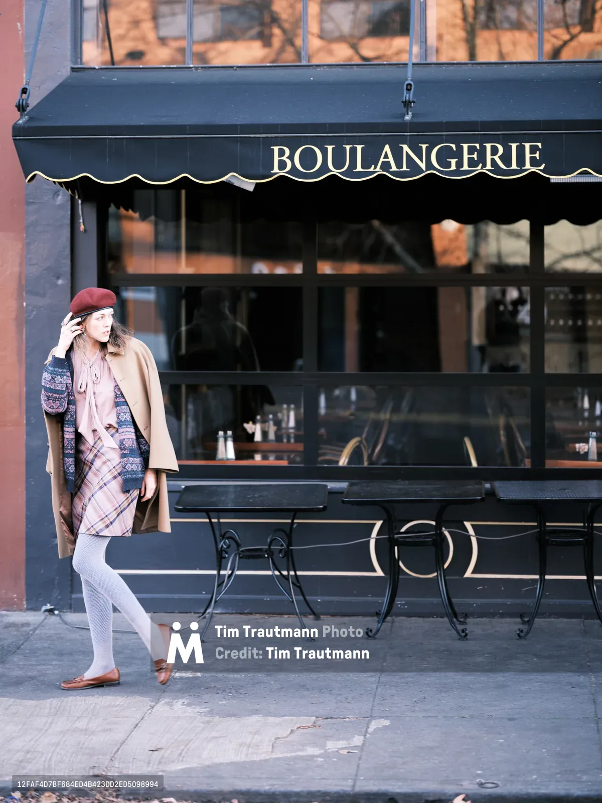 A young woman in a burgundy beret and plaid dress strolls past a charming boulangerie on Portland's Alberta Street, her layered autumn ensemble catching the warm afternoon light. The dark navy storefront with its classic French awning and wrought iron bistro tables creates an intimate European atmosphere against the urban Pacific Northwest backdrop. Her thoughtful pose, with one hand adjusting her beret, captures a moment of quiet contemplation as shadows dance across the sidewalk.