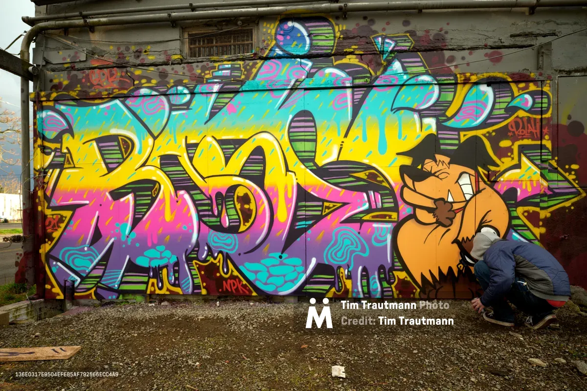 A graffiti artist crouches intently beside an explosive mural of electric blues, magentas, and yellows that transforms a weathered concrete wall in Portland's Alberta neighborhood. The dynamic lettering bursts with cartoon-like energy, featuring dripping paint effects and bold three-dimensional styling. Scattered debris and industrial pipes frame this urban canvas, capturing the raw creative spirit of Northeast Portland's street art scene.