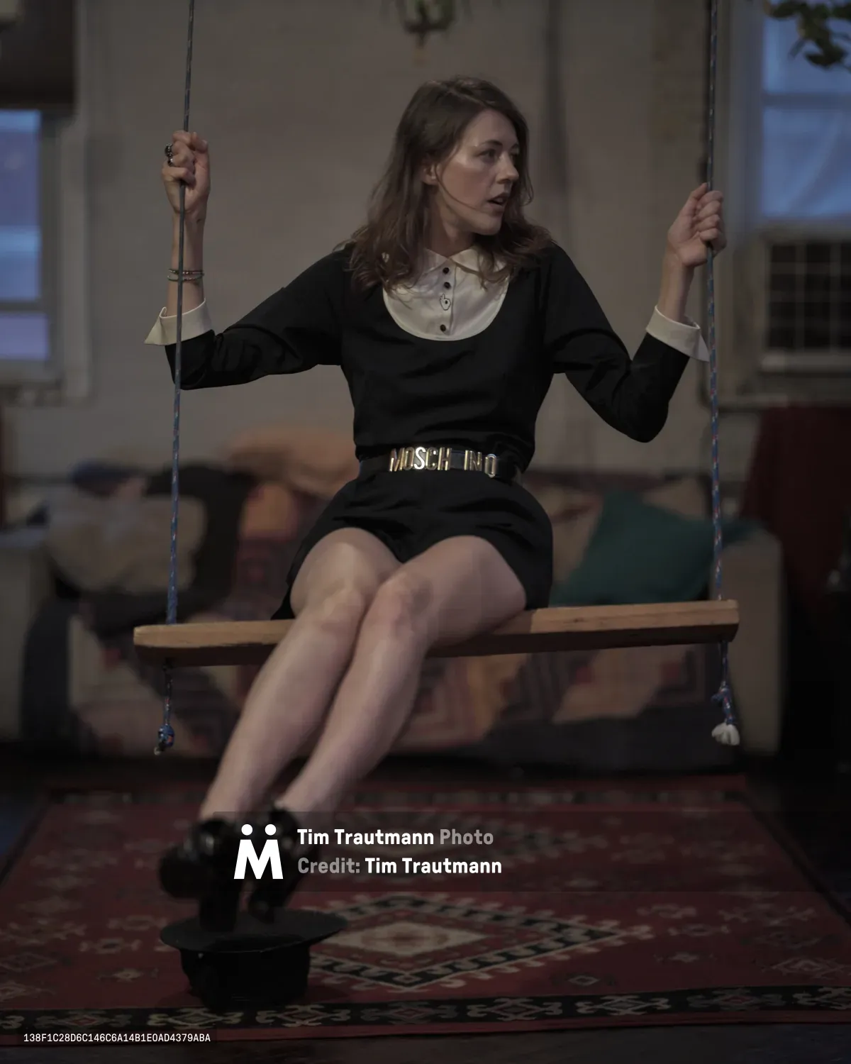 A young woman sits pensively on a wooden swing suspended in the warm interior of a Greenpoint apartment. Her dark dress contrasts with the cream-colored blouse beneath, while golden belt hardware catches the ambient light. The rope swing, weathered wooden seat, and patterned rug below create an intimate domestic tableau, with soft window light filtering through the Brooklyn space.