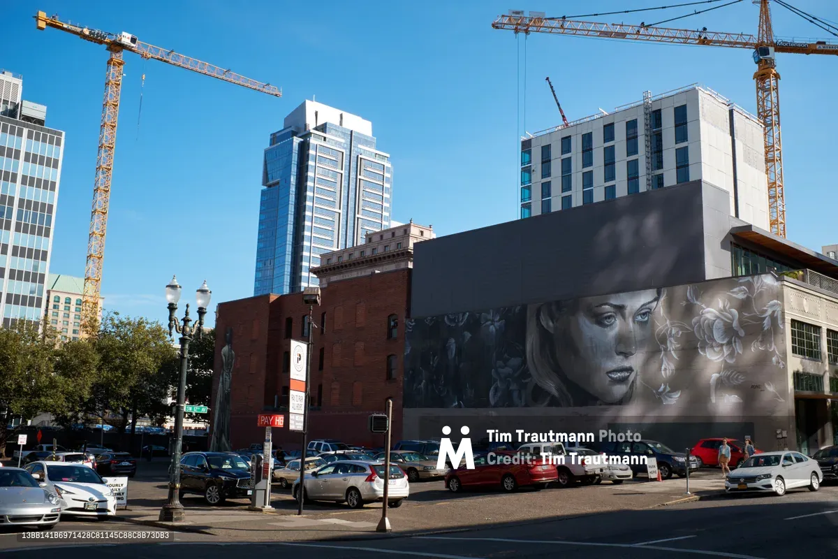 Two towering crane arms stretch across a brilliant blue sky above downtown Portland's Southwest Washington Street, where massive street murals transform the urban landscape below. A photorealistic portrait of a woman with flowing blonde hair dominates the right wall by artist RONE, while abstract geometric patterns in muted tones grace the left building by South African artist Faith47. The scene captures the intersection of development and artistic expression, with rows of parked cars creating foreground depth against the backdrop of gleaming glass towers and historic brick architecture.