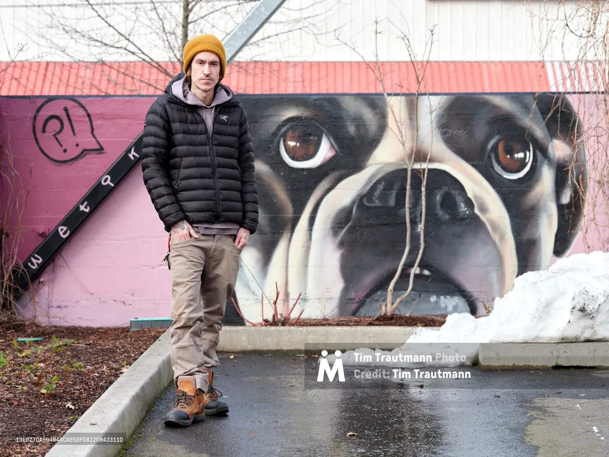 Artist Wetiko stands confidently beside his striking large-scale pug mural at Clay Creative Building in Southeast Portland's Hosford-Abernethy neighborhood. The young muralist, wrapped in a black puffer jacket and golden beanie against the winter chill, poses next to his hyperrealistic canine portrait that dominates the pink and coral building facade. Bare tree branches frame the scene while patches of snow dot the concrete landscape, capturing the intersection of urban art and Pacific Northwest winter.