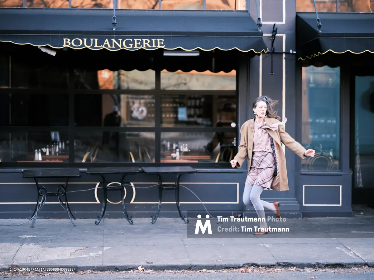 A young woman in motion hurries past a charming French bakery on Portland's Alberta Street, her camel coat and plaid dress flowing with the breeze. The classic navy blue storefront with traditional awning and wrought iron chairs creates an intimate European café atmosphere. Warm afternoon light illuminates the scene as she strides purposefully along the sidewalk, embodying the energy of urban life in this trendy neighborhood.