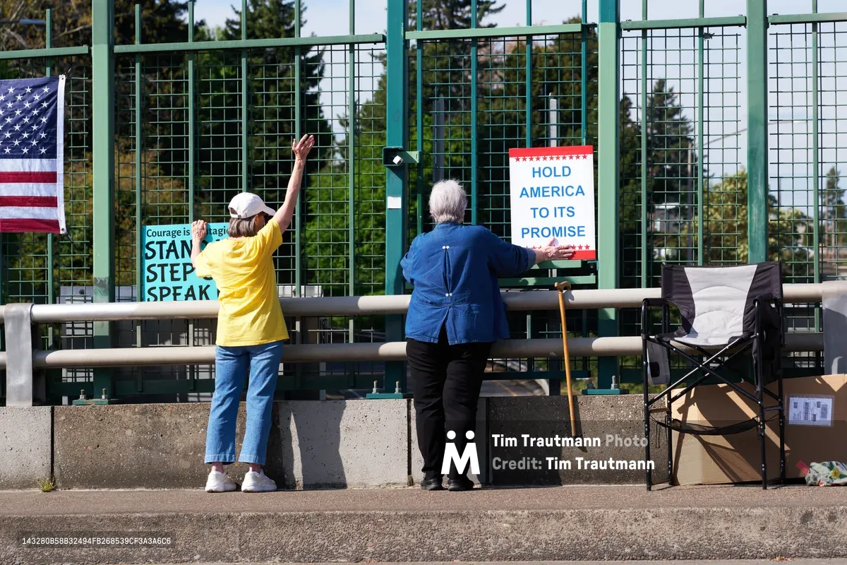 Two older activists prepare protest signage along the North Portland overpass above Interstate 5, their weathered hands gripping messages of resistance against the chain-link barrier. Morning light filters through the industrial mesh fencing as one woman in a bright yellow shirt raises her arm while her companion in denim secures a "Hold America to Its Promise" banner. Their determined silhouettes frame a moment of grassroots activism in the shadow of the Humboldt neighborhood's concrete infrastructure.