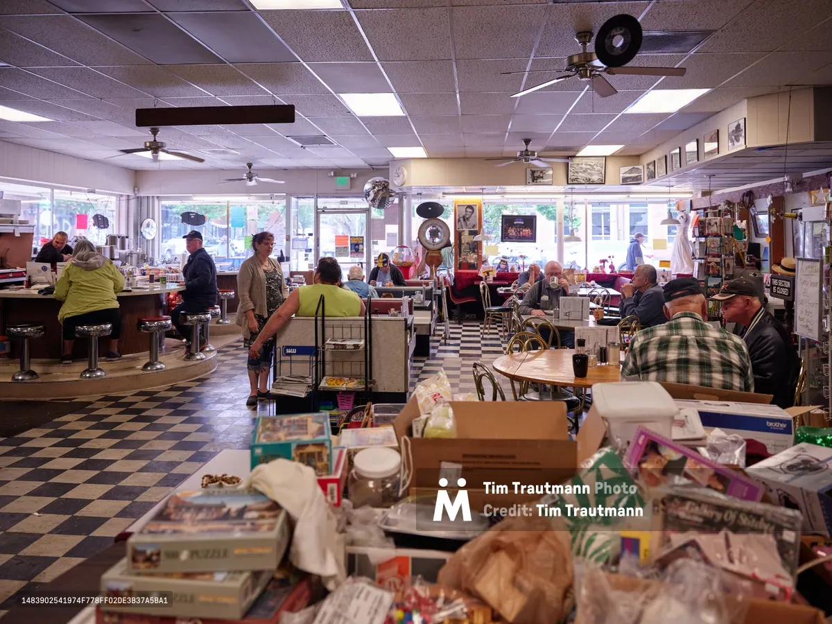 The warm interior of Pattie's Home Plate Café in Portland's Saint Johns neighborhood bustles with local patrons enjoying breakfast and coffee. Fluorescent lighting illuminates the classic diner atmosphere where seniors in flannel and casual wear occupy red vinyl booths and counter stools. Stacks of newspapers and magazines create an intimate foreground, while vinyl records and vintage photographs adorning the walls speak to the café's role as a cherished community gathering place.
