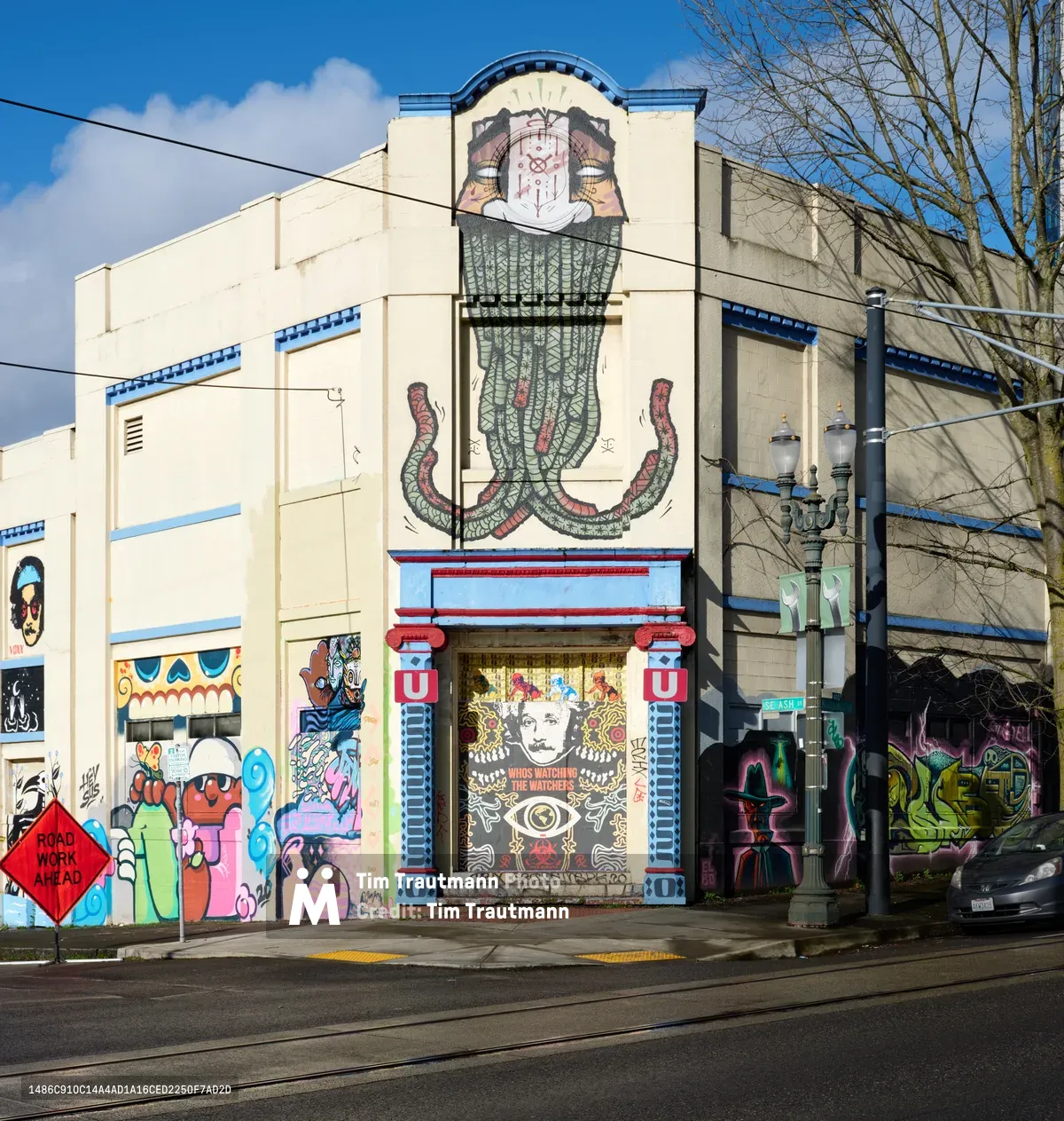 Vibrant street art adorns the cream-colored facade of a former Salvation Army warehouse in Southeast Portland's Central Eastside district. The expansive mural features a towering skeletal figure with ornate tentacles, flanked by colorful cartoon characters and intricate designs that transform the utilitarian building into an outdoor gallery. Power lines cross the scene under a partly cloudy sky, while bare winter trees frame the composition.