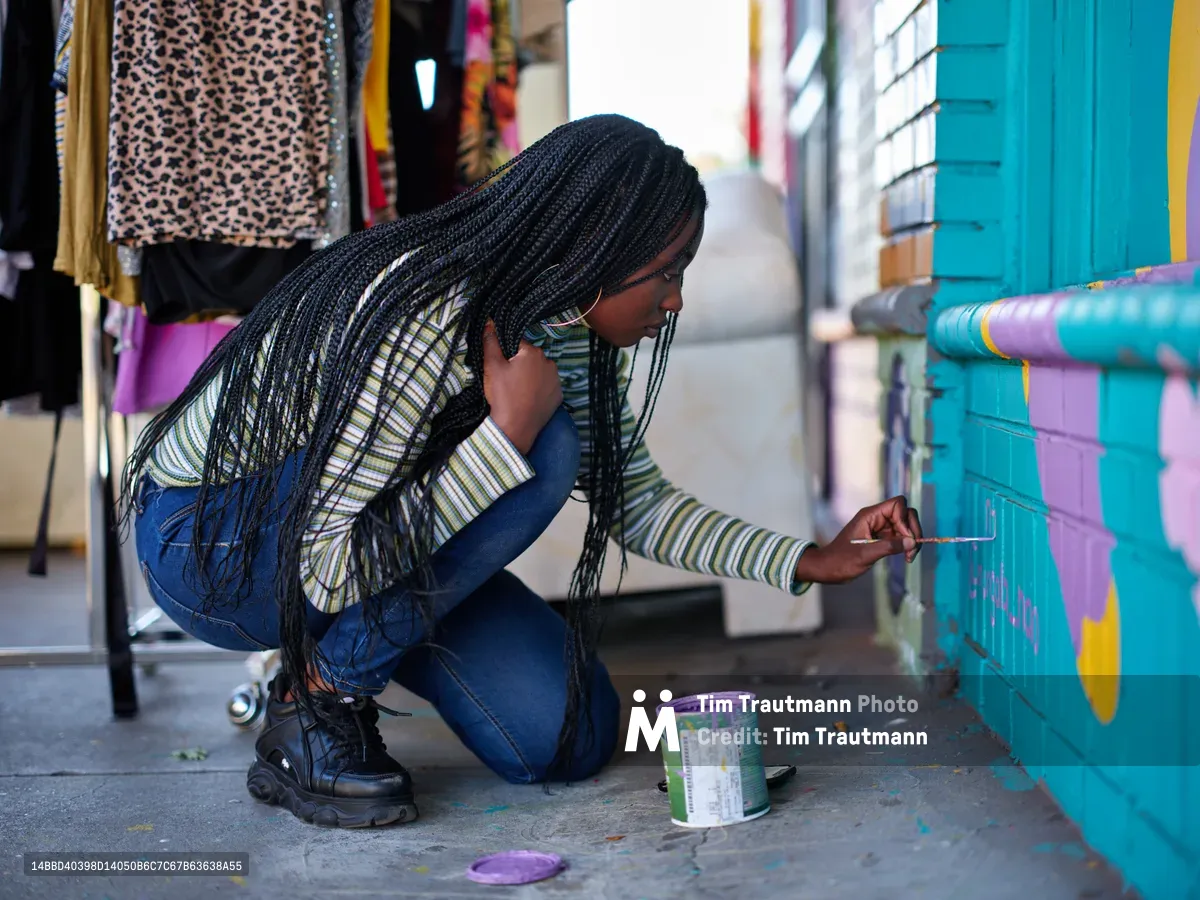 Naomi Likayi crouches in concentrated focus as she delicately signs her completed mural on a turquoise wall in Portland's vibrant Alberta Arts District. Her long black braids cascade forward as she works, paintbrush in hand, with a can of purple paint at her side. The urban street scene behind her reveals clothing displays and colorful storefronts, capturing the creative energy that defines this Northeast Portland neighborhood.