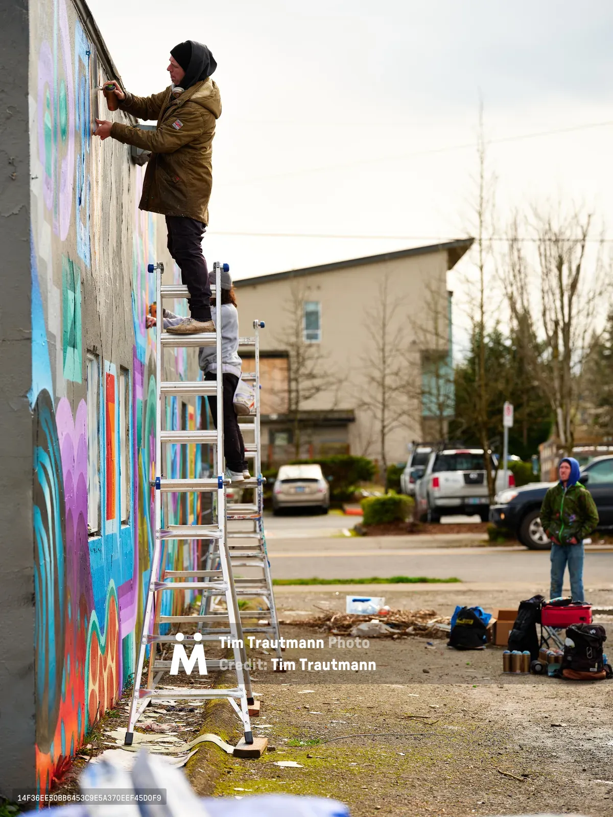 A hooded artist in an olive-green jacket stands on an aluminum ladder, spray painting a vibrant mural across a concrete wall in Northeast Portland, Oregon. The kaleidoscope of blues, purples, and warm tones transforms the urban facade while another observer watches from the sidewalk. Scattered art supplies and cans dot the pavement below, capturing the authentic process of community street art creation on an overcast day.