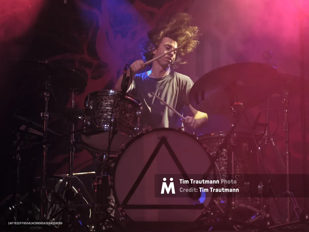 Julian Dorio, drummer for Band of Skulls, unleashes raw energy behind his kit during the band's 10 Year Anniversary Tour at Manhattan's legendary Webster Hall. His wild curls catch the intense stage lighting as drumsticks blur in motion, bathed in deep magenta and electric blue hues. The atmospheric concert photography captures the visceral power of live performance, with the drummer's focused expression illuminated against the moody backdrop of chrome hardware and shadowy venue architecture.