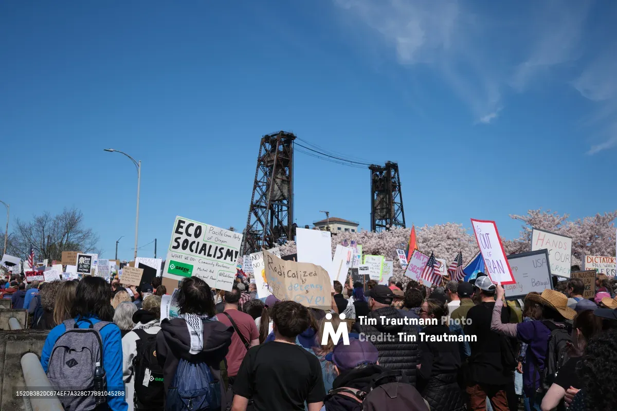 A diverse crowd of protesters fills the concrete steps along Northwest Naito Parkway in Portland's Old Town, their handmade signs creating a tapestry of dissent against the azure spring sky. The iconic Steel Bridge's weathered towers loom behind the demonstration, while delicate cherry blossoms frame the scene in pale pink contrast to the industrial backdrop. Protesters of all ages clutch homemade placards declaring messages like "Eco Socialism" and "No Kings," their collective voices rising beneath the bridge's steel latticework in this riverside gathering of democratic resistance.