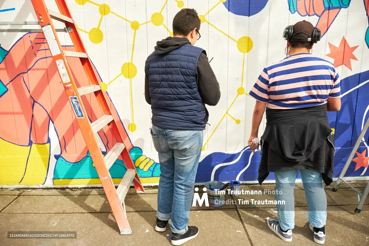 Two artists work intently on a vibrant public mural titled "Let's Talk" at Open Signal in Portland's historic Eliot neighborhood. The scene captures the collaborative creative process as Anke Gladnick and Maria Rodriguez paint geometric patterns and bold colors across the white wall. An orange ladder stands ready while paint buckets rest on the concrete sidewalk, with the emerging artwork featuring a dynamic interplay of yellow nodes, blue elements, and coral-toned forms that speak to community connection and dialogue.