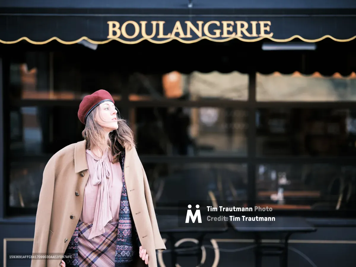 A young woman in a burgundy beret and camel wool coat pauses thoughtfully outside a traditional French bakery on Northeast Alberta Street in Portland's vibrant Alberta Arts District. Her layered outfit of soft pink blouse and patterned cardigan catches the gentle afternoon light as she gazes upward with a serene expression. The classic boulangerie storefront with its gold lettering and dark green awning creates an authentic European atmosphere against the urban Portland backdrop.