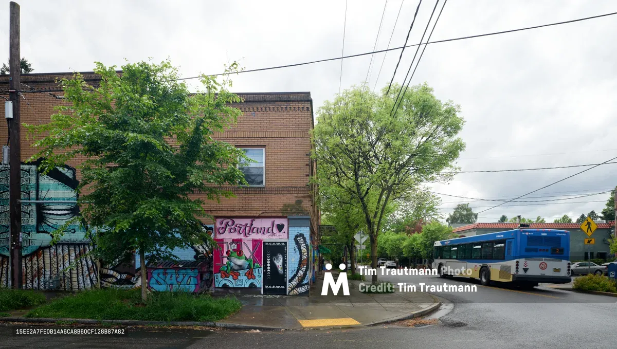 Vibrant street murals adorn the brick facade of the former Oregon Theater site on Southeast Division Street in Portland's Richmond neighborhood. The colorful artwork, featuring pink cursive lettering spelling 'Portland' alongside abstract graffiti elements, breathes new life into the weathered brick building under an overcast Pacific Northwest sky. A blue TriMet bus navigates the wet pavement, while power lines crisscross above the tree-lined residential street, capturing the authentic character of this evolving Portland neighborhood.