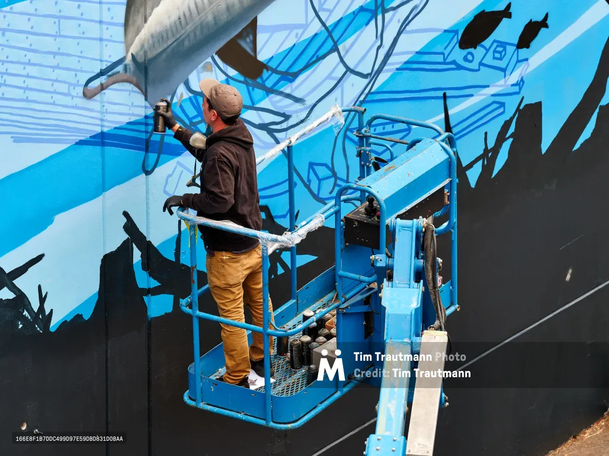 Street artist Jeremy Nichols commands a blue scissor lift against the stark concrete wall of Southeast Portland's Central Eastside district, his spray can releasing a cascade of azure tones across an emerging large-scale mural. Golden afternoon light bathes the industrial corridor of 2nd Avenue as the artist, clad in mustard work pants and brown hoodie, transforms the urban landscape into a flowing dreamscape of cerulean waves and abstract forms. The mechanical precision of the lift contrasts beautifully with the organic fluidity of his artistic vision, capturing the intersection of industrial infrastructure and creative expression that defines Portland's vibrant street art scene.