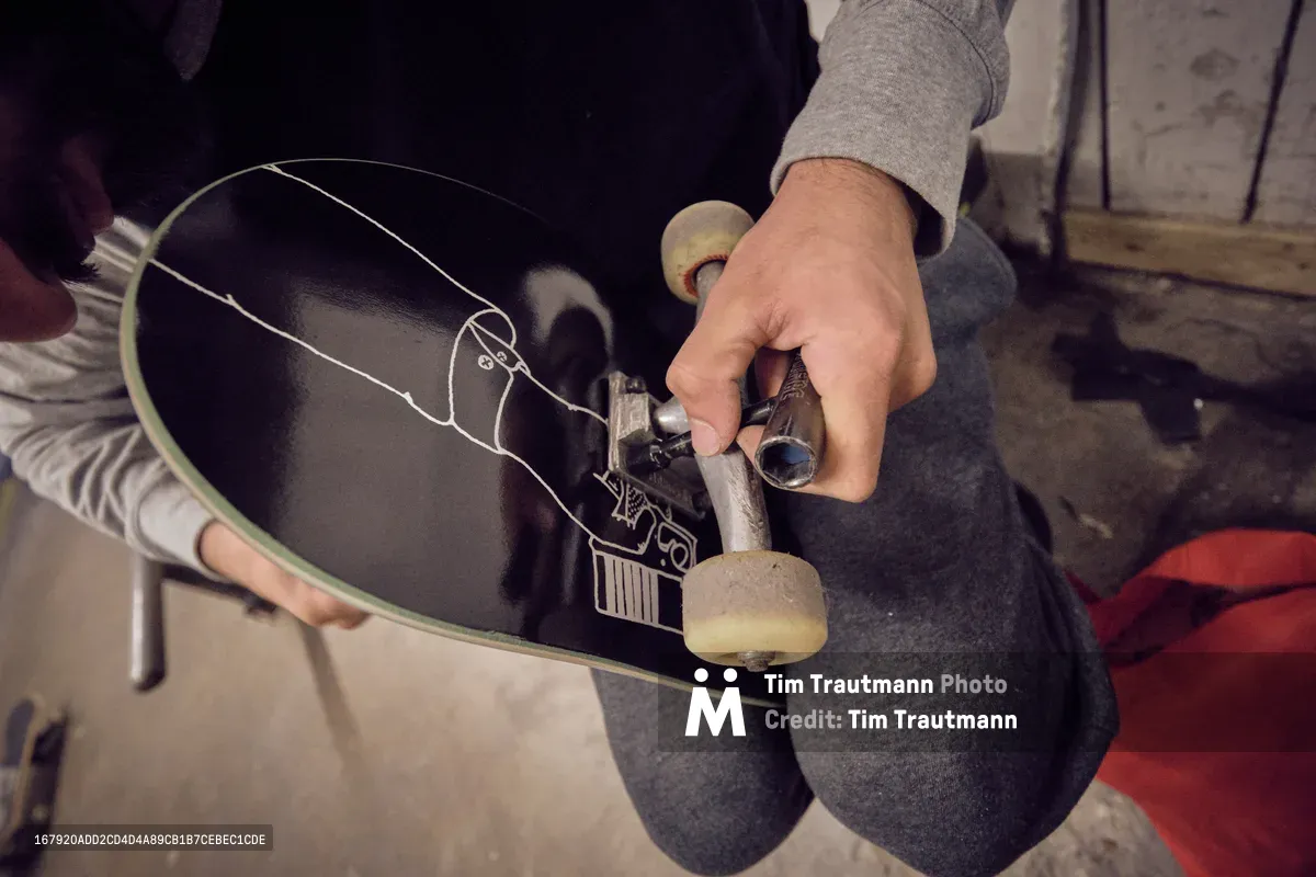 In the workshop atmosphere of Commonwealth Skateboarding on Southeast 20th Avenue in Portland's Buckman neighborhood, skilled hands make precise adjustments to a black skateboard's truck assembly. The intimate overhead perspective captures the mechanical intimacy of skate maintenance, with weathered concrete floors and workshop tools creating an authentic backdrop. Warm, diffused lighting bathes the scene in golden tones, emphasizing the tactile relationship between craftsperson and equipment in this celebrated Portland skate shop.