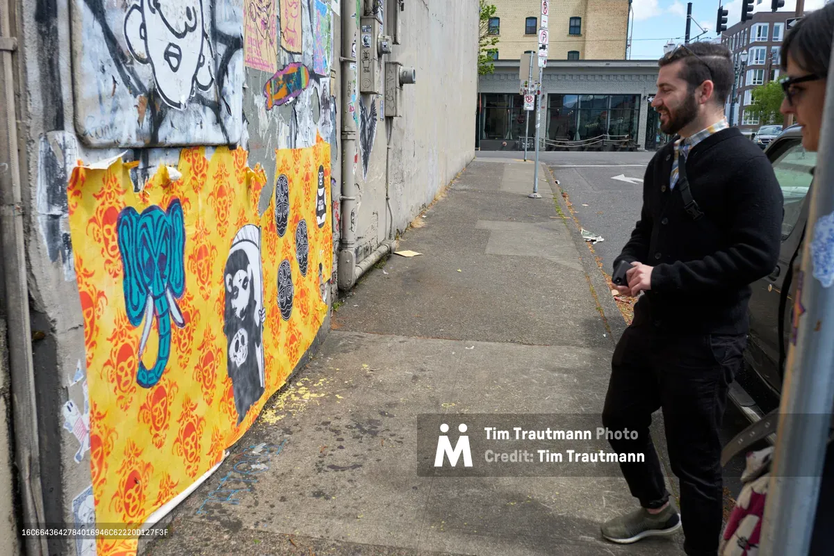 Tomás Valladares of the Portland Street Art Alliance guides participants through the vibrant wheat paste murals adorning concrete walls along Southeast Yamhill Street. Two men examine the layered urban artwork featuring bold yellow damask patterns, blue elephant imagery, and monochromatic character studies that transform the industrial corridor. The overcast Pacific Northwest light creates a muted atmosphere as the group moves through this celebrated gallery of sanctioned street art in Portland's evolving Central Eastside district.