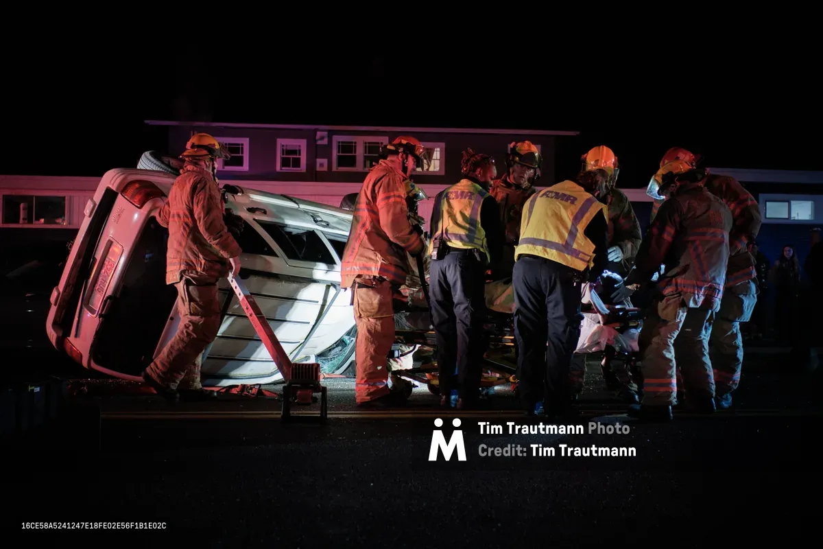 Portland firefighters and paramedics work urgently around an overturned silver SUV on Northeast Killingsworth Street in the Humboldt neighborhood. The scene is bathed in dramatic red and blue emergency lighting against the stark black night, creating an intense atmosphere of coordinated rescue efforts. Multiple first responders in high-visibility gear and protective helmets cluster around the damaged vehicle, their movements deliberate and focused as they tend to injured passengers. The residential backdrop of modest homes provides a sobering contrast to the chaos of twisted metal and emergency response.