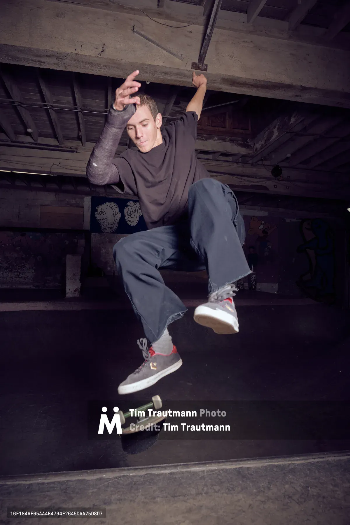 A young skateboarder hovers in mid-air beneath the weathered wooden beams of Commonwealth Skateboarding in Portland's Buckman neighborhood, his body frozen in graceful suspension above his board. The dramatic lighting cuts through the shadows of this basement skate haven, illuminating his focused expression and relaxed posture against the raw industrial architecture. The composition captures the poetry of skateboarding culture—where gravity momentarily surrenders to skill and style.