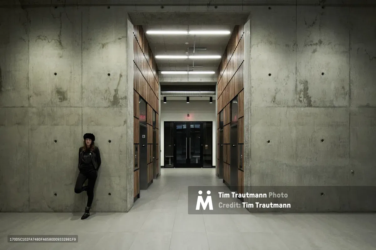 A woman in dark clothing and a black beanie leans against raw concrete walls in a stark modernist lobby space. The Pearl District building features towering concrete walls punctured by circular tie holes, while warm wood-paneled storage units create a striking contrast down the central corridor. Fluorescent lighting illuminates the space with clinical precision, emphasizing the brutal architectural geometry and the subject's contemplative solitude.