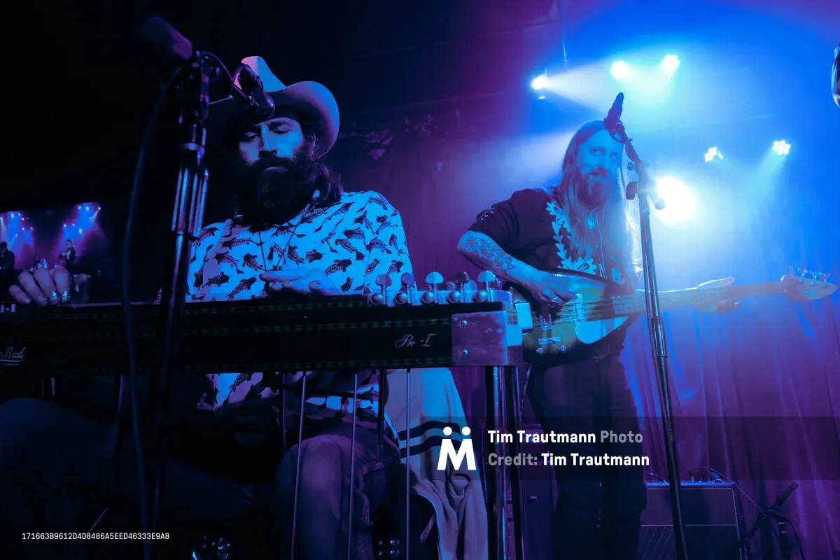 Barry Walker and Tobias Berblinger of Roselit Bone command the intimate stage at Dante's in Portland, bathed in ethereal blue and purple stage lighting. Walker, sporting a wide-brimmed hat and patterned shirt, works his synthesizer while Berblinger leans into his bass, both musicians caught in the transcendent moment of live performance. The moody lighting creates dramatic silhouettes against the smoky atmosphere of the legendary Portland venue.
