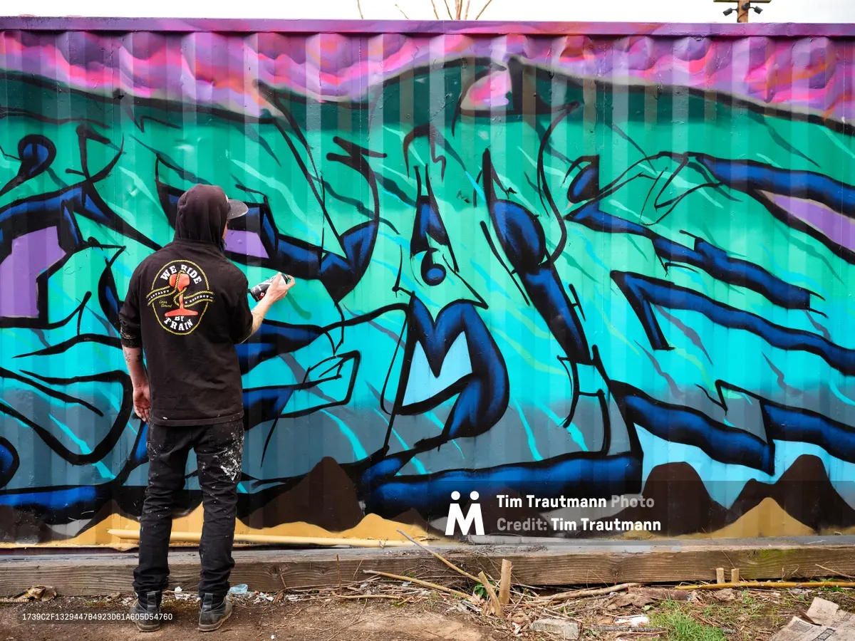 A street artist wearing a black hoodie and cap stands before an expansive graffiti mural, spray can in hand, adding details to the dynamic composition. The wall explodes with electric turquoise and deep blue wildstyle lettering, accented by magenta and purple geometric patterns that dance across the upper section. Set against the urban backdrop of Northeast Killingsworth Street in Portland's Alberta district, scattered debris and weathered concrete ground the scene in authentic street art culture.