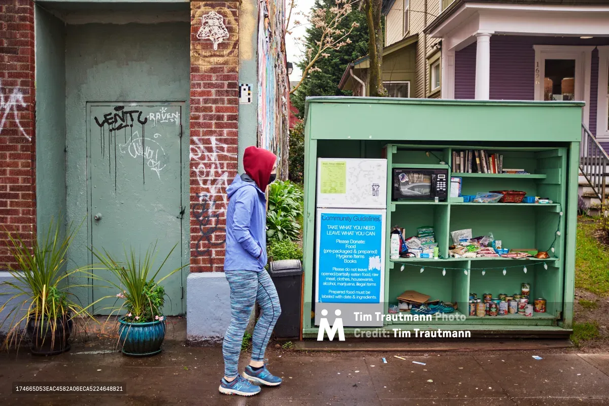 A person in a red beanie and blue hoodie approaches the PDX Free Fridge, a mint-green community pantry stocked with books, canned goods, and supplies along Alberta Street in Northeast Portland. The weathered brick building behind displays graffiti tags, while potted plants and residential homes create an intimate neighborhood setting. Soft overcast light captures this grassroots mutual aid initiative in action.