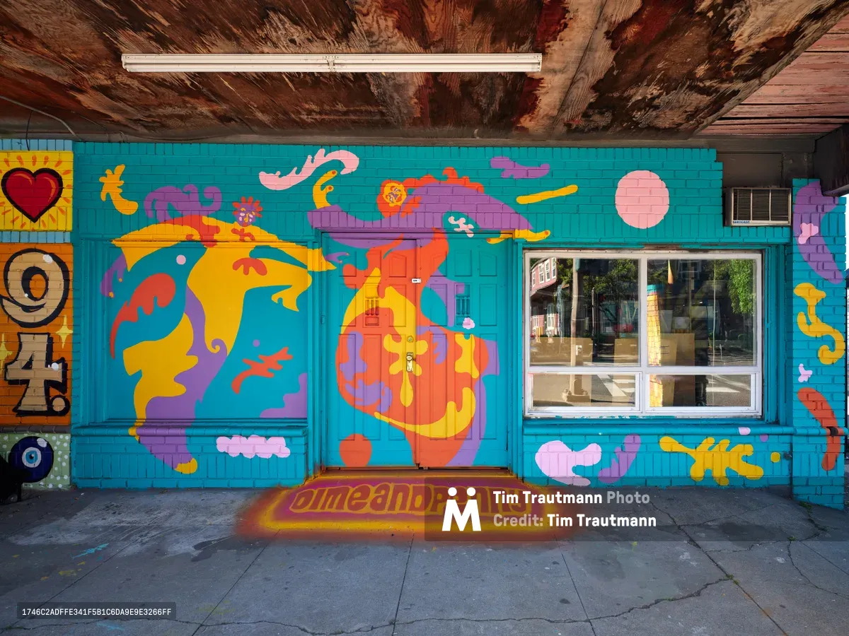 Artist Naomi Likayi's explosive mural transforms a turquoise wall in Portland's Alberta Arts District, featuring kaleidoscopic abstract human figures rendered in bold oranges, purples, and yellows against weathered wood ceiling beams. The psychedelic interpretation of flowing forms swirls with organic shapes and celestial elements, while "JimeAndPenny" appears stenciled on the concrete floor below. Natural light streams through the storefront window, illuminating this convergence of street art and community space in Northeast Portland's cultural corridor.