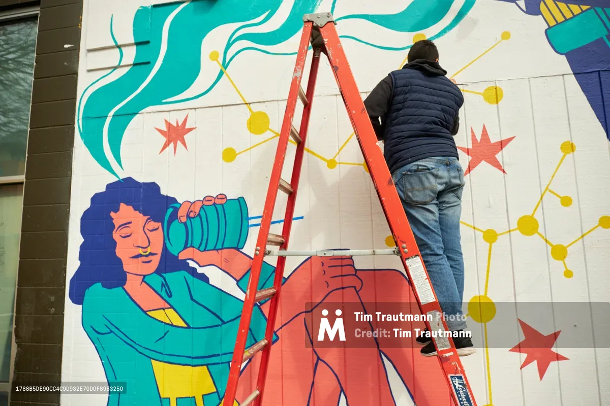 Artist Anke Gladnick climbs a red aluminum ladder to reach the upper portions of her vibrant mural "Let's Talk" on the exterior wall of Open Signal in Portland's Eliot neighborhood. The work-in-progress reveals her signature style of layered analog and digital elements, featuring flowing turquoise forms, geometric constellations of yellow dots, coral-pink star bursts, and a serene female figure with dark curls holding what appears to be a green water vessel. Gladnick, dressed in a black puffer jacket and jeans, stretches upward with concentrated focus, embodying the physical dedication required to transform architectural surfaces into dreamlike narratives.